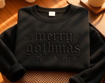 May include: Black sweatshirt with the phrase "merry gothmas" in gothic lettering, accented with snowflakes. The crew neck sweatshirt is folded on a chair, with a coffee cup in the background.