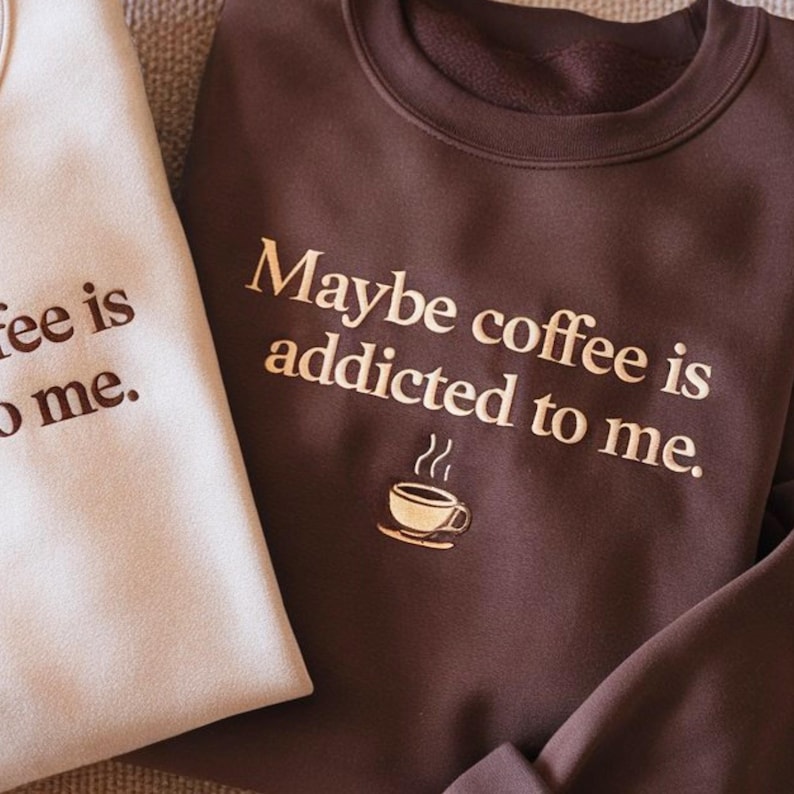 May include: A dark brown crewneck sweatshirt featuring the gold text "Maybe coffee is addicted to me." and a small gold coffee cup graphic. Beside it, a portion of a light beige sweatshirt is visible with matching brown text.
