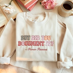 Könnte beinhalten: Cremefarbenes Sweatshirt mit dem gestickten Text "BUT DID YOU DOCUMENT IT?" in Retro-Schrift. Darunter steht "- Human Resources" in kleiner, kursiver Schrift. Das Sweatshirt liegt gefaltet auf einer weißen Oberfläche.