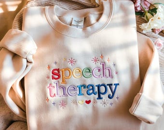 May include: A cream-coloured sweatshirt embroidered with the words "speech therapy" in rainbow colours. The design includes stars and hearts. The sweatshirt has a crew neck and long sleeves. The garment is displayed on a neutral-toned surface.