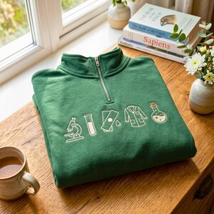 Peut inclure: Un sweat-shirt vert forêt à fermeture éclair quart de longueur, orné de broderies blanches représentant un microscope, un tube à essai, une lame de microscope, une blouse de laboratoire et un ballon à fond rond sur la poitrine.