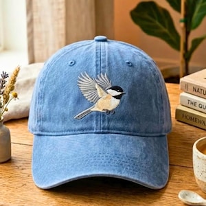 May include: A light blue denim baseball cap featuring an embroidered design of a small bird with a black head, white cheeks, and tan wings in flight. The cap has a structured crown, a curved brim, and visible stitching across the front panels.