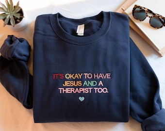 May include: Navy blue sweatshirt with the embroidered text "IT'S OKAY TO HAVE JESUS AND A THERAPIST TOO." The text is in various colors, with a small blue heart below. The sweatshirt is folded, with the sleeves visible.
