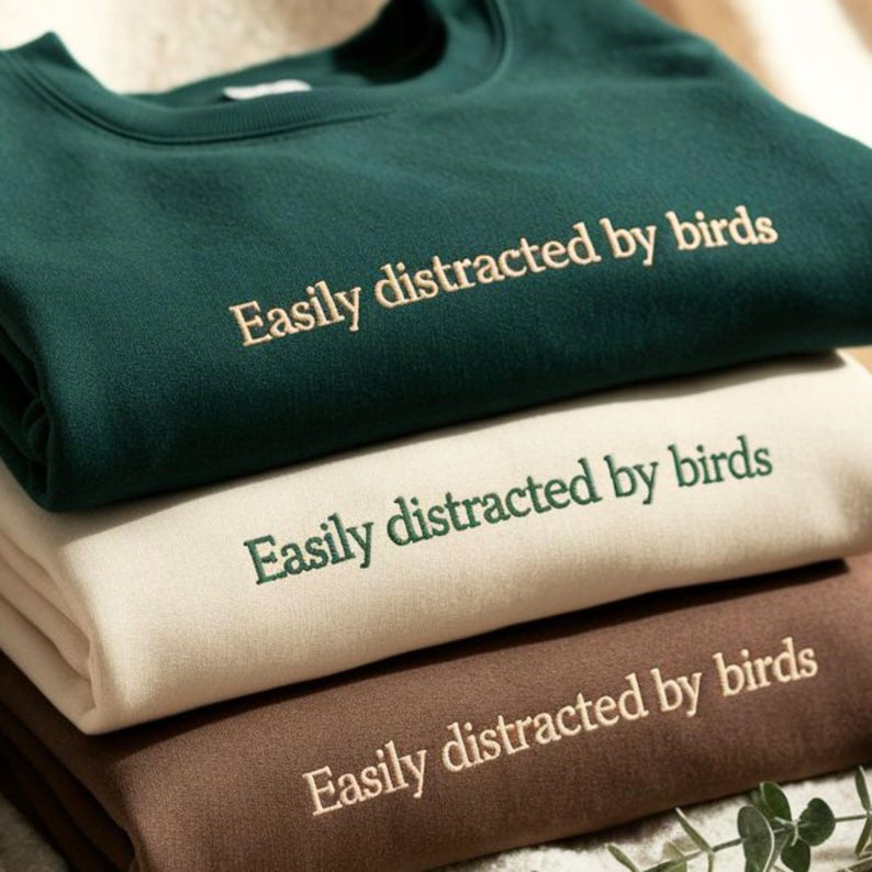 May include: A stack of three crewneck sweatshirts in dark green, cream, and brown. Each sweatshirt features the embroidered text "Easily distracted by birds" centered on the chest. The top sweatshirt has cream text, the middle has green, and the bottom has cream.