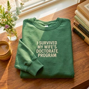 May include: A green sweatshirt with the text "I SURVIVED MY WIFE'S DOCTORATE PROGRAM" in white. The sweatshirt is folded on a wooden surface, with a cup of coffee and a vase of flowers nearby.