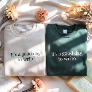 May include: Two crewneck sweatshirts, one beige with dark green embroidered text and one dark green with beige embroidered text. Both feature the phrase 'it's a good day to write' centered on the chest in a simple, clean, lowercase font.