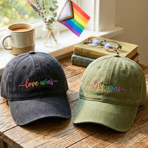May include: Two baseball caps with the embroidered phrase "love wins" in rainbow colors. One cap is dark gray, the other is olive green. A Pride flag and other items are in the background.