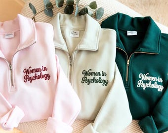 May include: Three quarter-zip sweatshirts in pink, beige, and dark green. Each sweatshirt has the embroidered text "Women in Psychology" on the front. The sweatshirts are displayed on a cream-coloured surface.