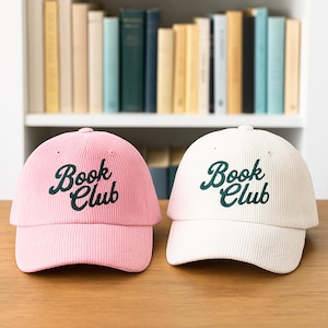 May include: Two corduroy baseball caps, one pink and one white, with the words "Book Club" embroidered in dark green. The caps are displayed on a wooden surface with a bookshelf in the background.