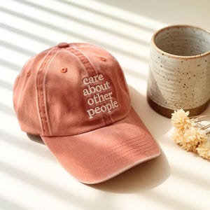 May include: A dusty rose baseball cap with the embroidered text "Care About Other People" in white. The cap is next to a speckled ceramic mug and a small bundle of dried flowers.