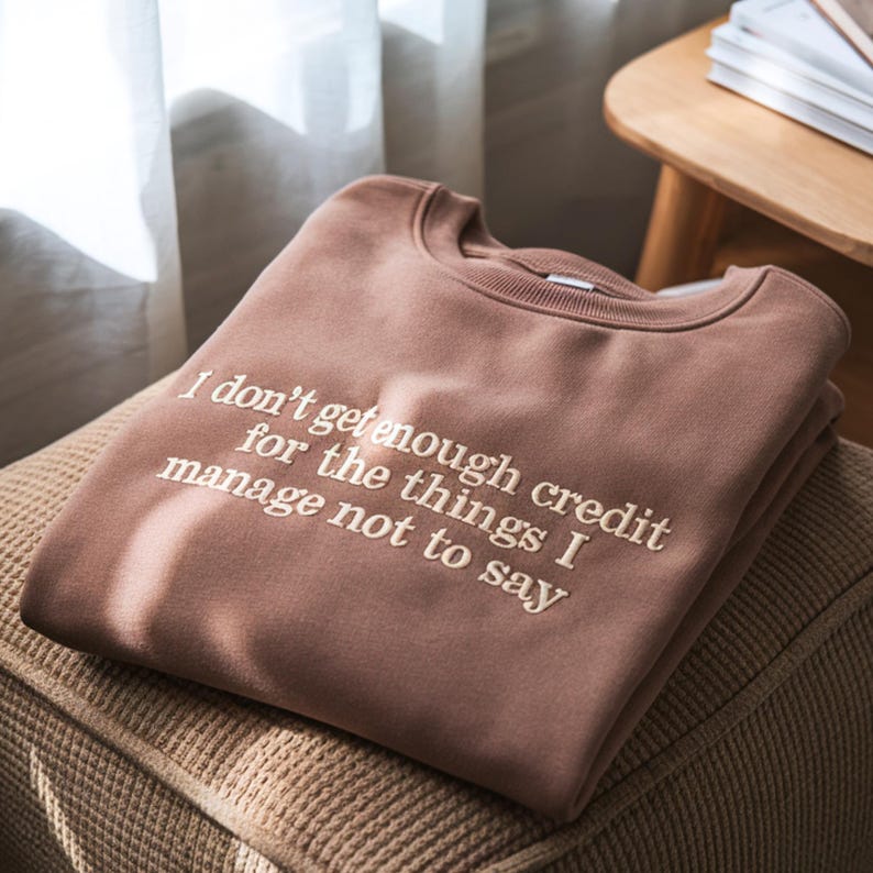 May include: A brown sweatshirt with the embroidered text "I don't get enough credit for the things I manage not to say." The sweatshirt is folded and resting on a textured surface. The image is well-lit, showcasing the garment's details.