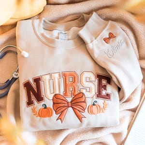 May include: A beige crewneck sweatshirt featuring the word NURSE embroidered in brown and cream varsity-style letters. The design includes an orange bow and two pumpkins. The sleeve is embroidered with a small orange bow and the name Olivia.