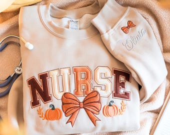 May include: A beige sweatshirt with the word "NURSE" embroidered in brown and cream-colored letters. The design includes an orange bow, pumpkins, and the name "Olivia" embroidered on the sleeve. A stethoscope is visible.