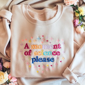 May include: Beige sweatshirt with embroidered text "A moment of science please" in colorful letters. The design includes embroidered stars, hearts, and other shapes in various colors. The sweatshirt is folded neatly.