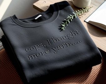 May include: A dark gray sweatshirt with the embroidered text "oops, I bought more yarn...." is displayed on a wooden surface. A mug, a sprig of greenery, and an open book are also visible.