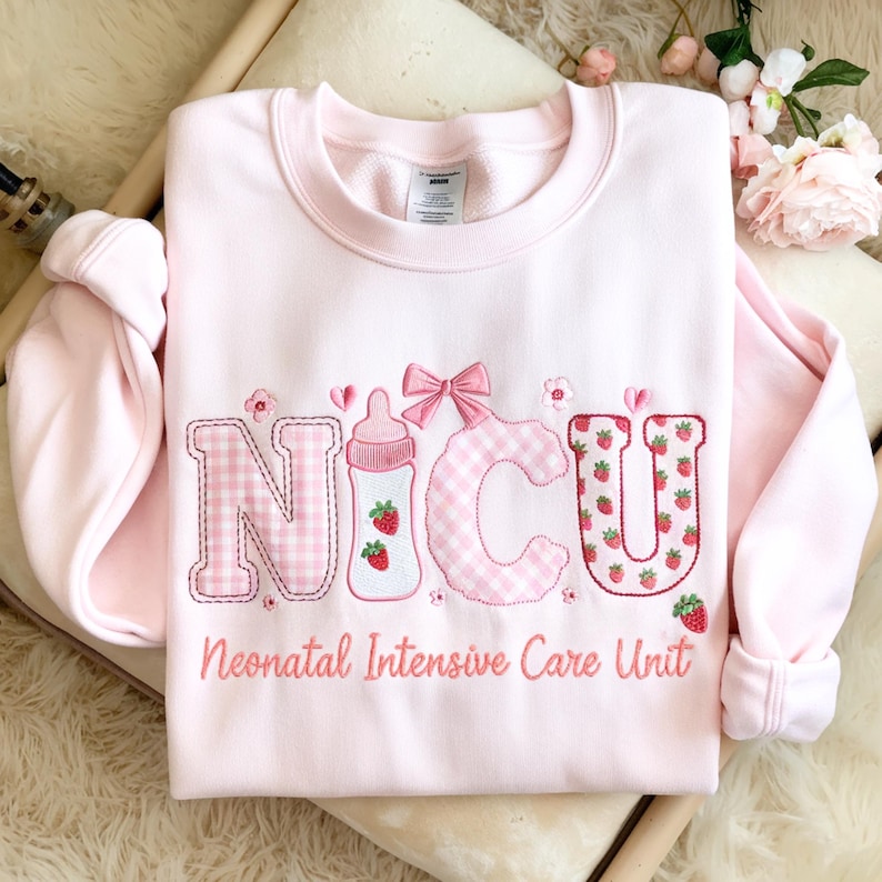 May include: A pale pink sweatshirt featuring the embroidered letters "NICU". The "N" is checkered, the "I" is a baby bottle with strawberries, the "C" is checkered, and the "U" is covered in strawberries. The words "Neonatal Intensive Care Unit" are embroidered below.