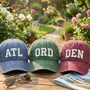 May include: Three baseball caps in blue, green, and burgundy, each with white embroidered letters spelling "ATL", "ORD", and "DEN". Sunglasses and a magazine are on the wooden surface in front of the caps. The background features a garden with flowers.