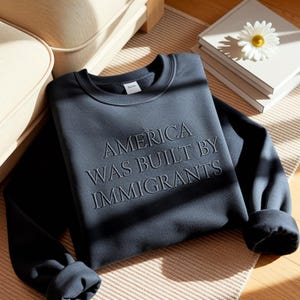 May include: A dark blue sweatshirt with the embroidered text "AMERICA WAS BUILT BY IMMIGRANTS." The crew neck sweatshirt is folded on a neutral-toned rug, with a stack of books and a daisy in the background.