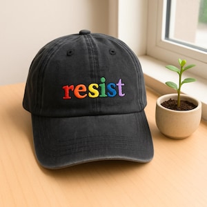 May include: A black baseball cap with the word "resist" embroidered in rainbow colours across the front. The cap is made of a soft, worn fabric and has a curved brim. The cap is sitting on a wooden surface.