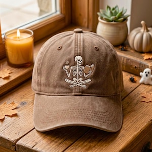 May include: A brown baseball cap featuring an embroidered graphic of a white skeleton sitting in a cross-legged position, holding a steaming cup of coffee in one hand and an open book in the other, centered on the front panel of the cap.