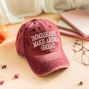 May include: A maroon baseball cap with the embroidered text "IMMIGRANTS MAKE AMERICA GREAT" in white. The cap has a distressed, vintage look, with visible stitching and a slightly worn brim. The background includes books and glasses.