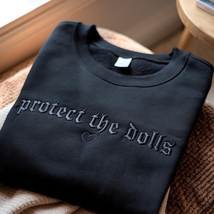 May include: Black crewneck sweatshirt with the embroidered phrase "protect the dolls" in gothic lettering, along with a small heart. The sweatshirt is folded and resting on a textured surface.