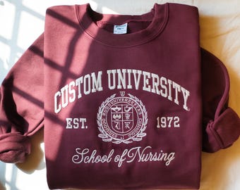 May include: A maroon sweatshirt with white embroidered text reading "Custom University" above a crest and "School of Nursing" below. The sweatshirt also has the text "EST. 1972". The sleeves are folded.