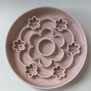 K&ouml;nnte beinhalten: Eine hellrosa Hunde-Slow-Feeder-Sch&uuml;ssel mit Blumendesign. Die Sch&uuml;ssel hat eine runde Form mit erh&ouml;htem Rand und bl&uuml;tenf&ouml;rmigen Hindernissen im Inneren, um das Fressen zu verlangsamen. Die Mitte der Sch&uuml;ssel hat einen kleinen kreisf&ouml;rmigen Bereich.