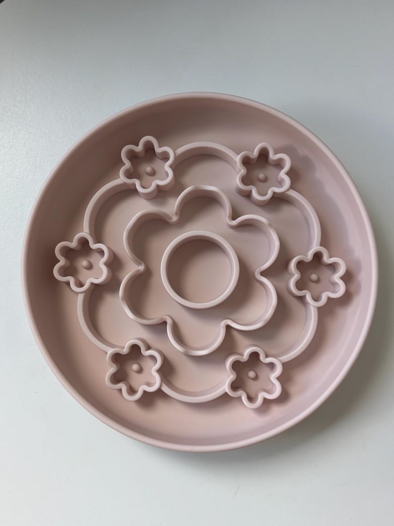 K&ouml;nnte beinhalten: Eine hellrosa Hunde-Slow-Feeder-Sch&uuml;ssel mit Blumendesign. Die Sch&uuml;ssel hat eine runde Form mit erh&ouml;htem Rand und bl&uuml;tenf&ouml;rmigen Hindernissen im Inneren, um das Fressen zu verlangsamen. Die Mitte der Sch&uuml;ssel hat einen kleinen kreisf&ouml;rmigen Bereich.