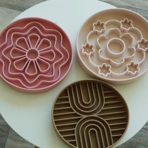 K&ouml;nnte beinhalten: Drei runde Futtern&auml;pfe f&uuml;r Haustiere in Rosa- und Braunt&ouml;nen. Die N&auml;pfe haben einzigartige Designs, darunter eine Blume und ein Regenbogenmuster. Die N&auml;pfe sind aus einem strapazierf&auml;higen Material gefertigt und sollen das Fressen verlangsamen.