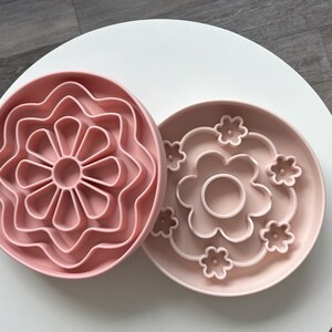 K&ouml;nnte beinhalten: Zwei rosa, bl&uuml;tenf&ouml;rmige Silikon-Slow-Feeder-Schalen. Eine Schale hat ein mehrbl&auml;ttriges Design, die andere einen Blumenumriss mit kleineren Blumenakzenten. Die Schalen sollen das Fressen von Haustieren verlangsamen.