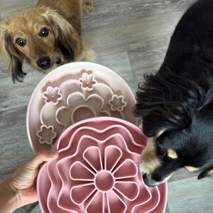 K&ouml;nnte beinhalten: Zwei Futtern&auml;pfe f&uuml;r Hunde mit langsamer F&uuml;tterung. Einer ist hellrosa mit einem Blumenmuster, der andere ist dunkler rosa mit einem Labyrinthmuster. Zwei Hunde schauen auf die Schalen.