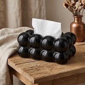 May include: A rectangular tissue box cover featuring a glossy black finish and a distinct bubble-like, spherical texture across its exterior. The box is designed with a central opening at the top, through which a single white tissue is pulled upward.