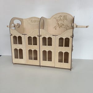 May include: A wooden model of a horse stable with the word "Stajnia" written on the front. The stable has multiple stalls with arched windows and a roof with a decorative design.