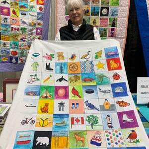 Puede incluir: Una colcha de tela blanca con una cuadrícula de 48 cuadrados, cada uno con una ilustración diferente pintada a mano de un icono canadiense. Las ilustraciones incluyen un oso, un alce, una hoja de arce, un castor, un disco de hockey, una canoa, un faro, un tomate, una abeja, un libro, un gato, un coche, un pájaro, un copo de nieve, una estrella, una mazorca de maíz, un pez, una mariquita, un dinosaurio, una ballena, un búfalo, un tipi, un globo, un cardenal, un par de calcetines, un arrendajo azul, una botella de jarabe de arce, una mariposa, un par de patines de hielo, un jugador de hockey, un árbol, un barco, una bandera canadiense, un vaso de agua, un castor y una cordillera. La colcha se exhibe en una mesa con un cartel que dice "Por favor, manipule con cuidado".