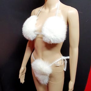May include: A white faux fur bikini set. The top has fluffy, round cups with thin straps. The bottom is a matching thong with a fur accent and tie closure. The set is displayed on a mannequin against a black background.