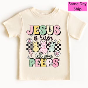 May include: Cream-colored t-shirt with the words "JESUS is risen tell your PEEPS" in colorful, retro-style lettering. The design includes three Easter bunny figures, lightning bolts, and flowers.