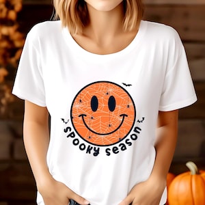 May include: White t-shirt with a Halloween design. The design features an orange smiley face with a spiderweb pattern, spiders, and bats. The words "Spooky Season" are written in a curved font around the smiley face.
