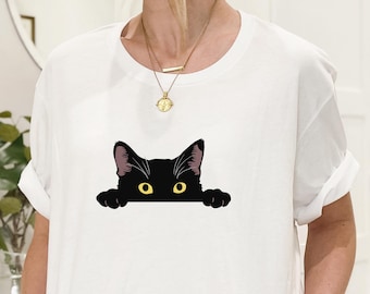 May include: White t-shirt featuring a black cat graphic. The cat has yellow eyes and is peeking over the bottom of the shirt. The shirt has short sleeves and a round neckline. A gold necklace is visible.