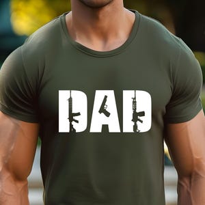 May include: Olive green t-shirt with the word "DAD" in white block letters. The letter "D" features a silhouette of a rifle, the "A" a handgun, and the second "D" a rifle. The shirt is made of a soft material.