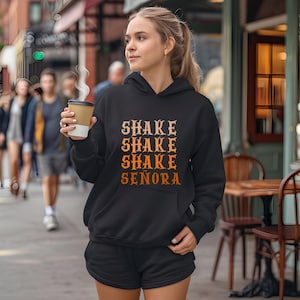 Op de afbeelding: Een zwarte hoodie met de tekst SHAKE SHAKE SHAKE SENORA in een verloop van oranje en gele tinten op de voorzijde. De trui heeft lange mouwen, een kangoeroezak aan de voorzijde en een bijpassende zwarte korte broek.