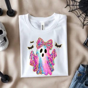 May include: White t-shirt featuring a colorful ghost design with a bow and bats. The ghost has a pink, blue, and yellow gradient with leopard print accents. The shirt is made of a soft material and is perfect for Halloween.