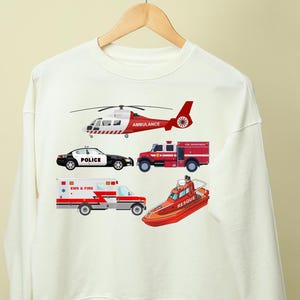 May include: White sweatshirt featuring illustrations of emergency vehicles: a red and white helicopter, a black and white police car, a red fire truck, an ambulance, and a rescue boat. The sweatshirt is hanging on a wooden hanger.