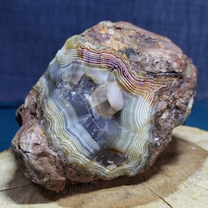 Puede incluir: Una geoda de ágata rugosa y sin pulir que presenta bandas concéntricas de colores blanco, gris y marrón rojizo. El centro de la piedra revela una cavidad cristalina translúcida con una formación de cristal clara, lisa y prominente.