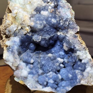 May include: A natural geode with a striking blue and white crystalline interior. The geode's exterior is a rough, earthy brown, while the inside features a cluster of small, rounded crystals. The geode is displayed on a wooden slice.