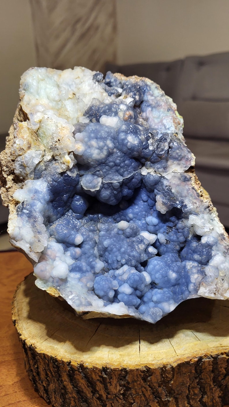 May include: A natural geode with a striking blue and white crystalline interior. The geode's exterior is a rough, earthy brown, while the inside features a cluster of small, rounded crystals. The geode is displayed on a wooden slice.