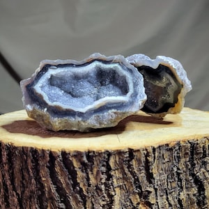 Puede incluir: Dos mitades de una geoda con un interior de cristal morado y blanco. La geoda está sentada sobre un tronco de madera.