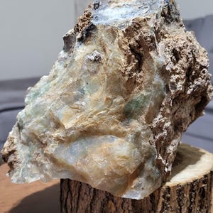 May include: A large, roughly shaped stone with a mix of tan, green, and white hues, displayed on a natural wood slice. The stone has a textured surface, and the wood slice provides a rustic aesthetic.