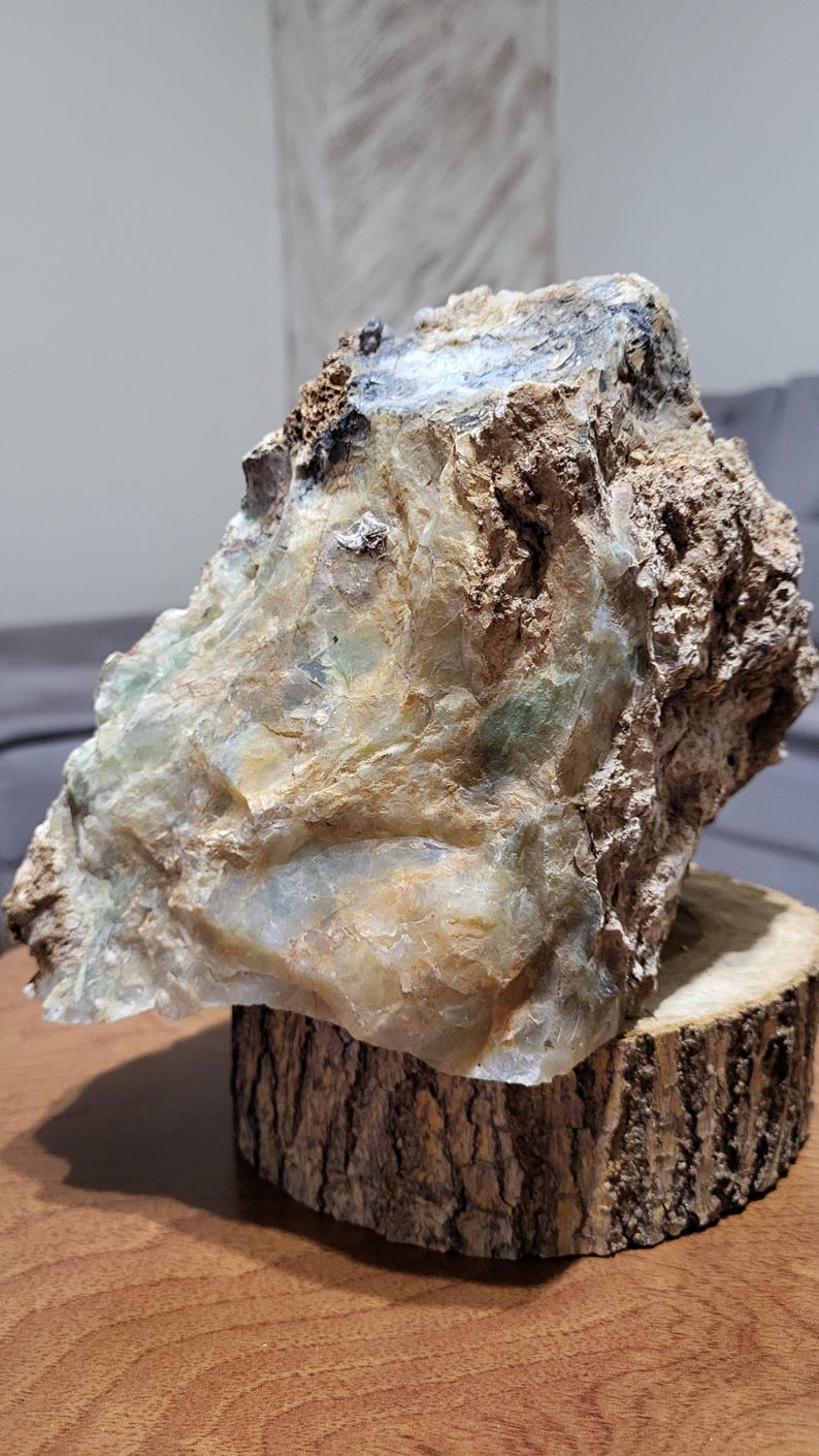 May include: A large, roughly shaped stone with a mix of tan, green, and white hues, displayed on a natural wood slice. The stone has a textured surface, and the wood slice provides a rustic aesthetic.