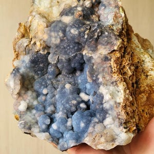 May include: A natural geode specimen with a cluster of blue chalcedony crystals. The geode has a rough, brown exterior and a cavity filled with small, rounded, blue-grey formations. The overall appearance is textured and organic.